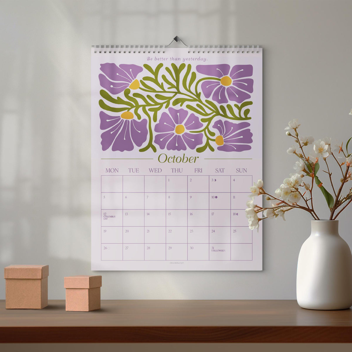 May include: A wall calendar for October with a floral design in purple and green. The calendar has a spiral binding and is hanging on a wall. The text "Be better than yesterday" is at the top.
