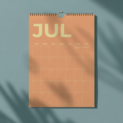 May include: A peach-colored wall calendar for July, with the month's name in large, light-yellow letters. The calendar grid is visible, with days of the week labeled across the top. A silver ring holds the calendar, which is hanging against a teal background.