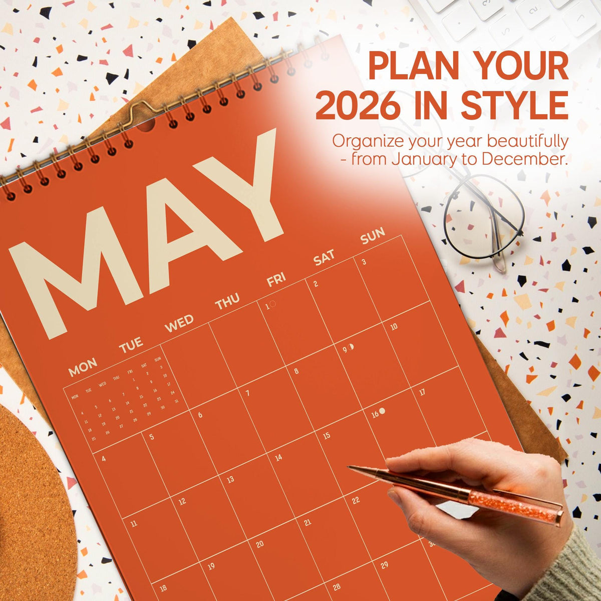 May include: An orange 2026 calendar open to May, with the text "PLAN YOUR 2026 IN STYLE". A hand holding a pen is pointing to a date on the calendar. The calendar is on a patterned surface.
