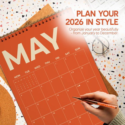 May include: An orange 2026 calendar open to May, with the text "PLAN YOUR 2026 IN STYLE". A hand holding a pen is pointing to a date on the calendar. The calendar is on a patterned surface.