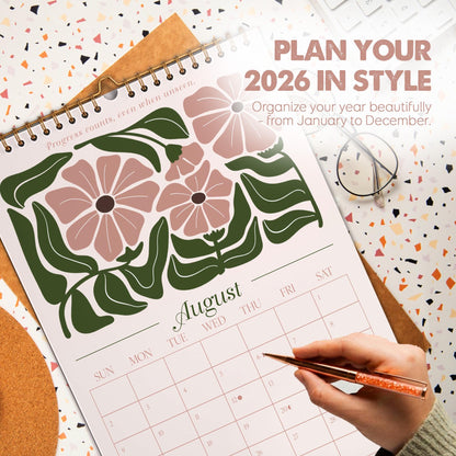 May include: A spiral-bound 2026 calendar with a floral design in pink and green. The calendar is open to August, with the text "Plan Your 2026 In Style" at the top. A hand is holding a pen, pointing to a date.