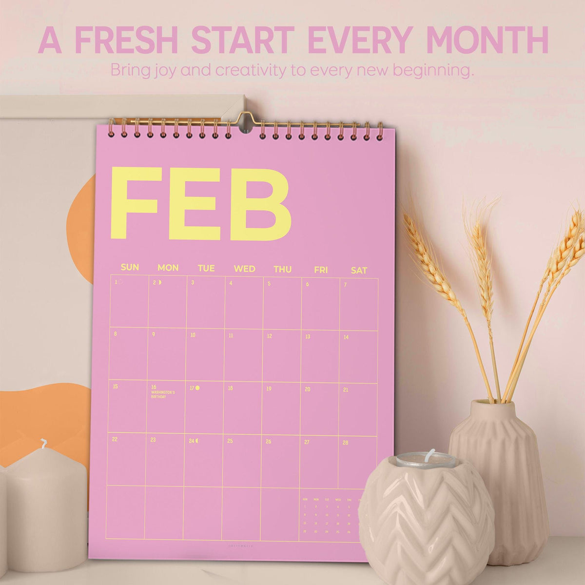 May include: A pink wall calendar for February with yellow text. The calendar has a spiral binding and is hanging on a wall. The text at the top says "A FRESH START EVERY MONTH". There are decorative items in the foreground.