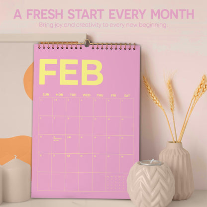May include: A pink wall calendar for February with yellow text. The calendar has a spiral binding and is hanging on a wall. The text at the top says "A FRESH START EVERY MONTH". There are decorative items in the foreground.