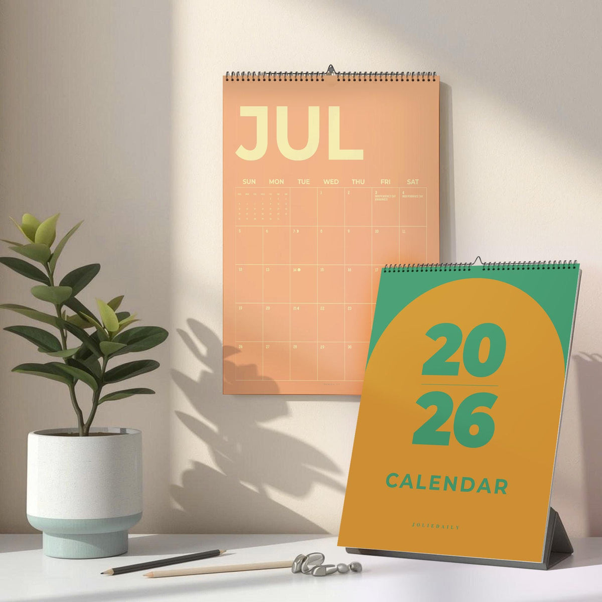 May include: Two calendars, one hanging and one standing, are displayed. The hanging calendar is peach-colored and shows the month of July. The standing calendar is green and yellow, with the year 2026 and the word "CALENDAR" printed on it.