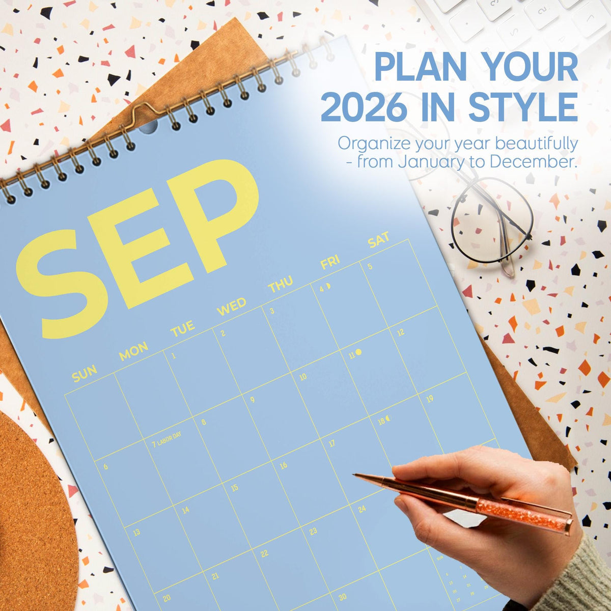 May include: A light blue 2026 calendar open to September, with yellow lettering. The calendar is on a brown backing with a gold spiral binding. Text reads "PLAN YOUR 2026 IN STYLE". A hand holds a rose gold pen.