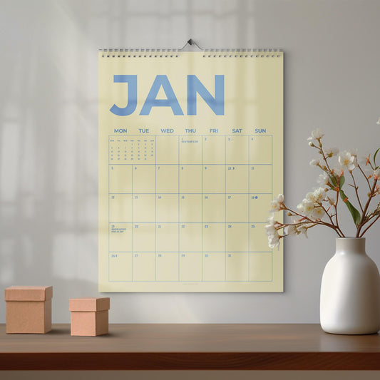 May include: A wall calendar for January with a cream background and blue text. The word "JAN" is at the top, with the days of the week below. A white vase with white flowers and two small brown boxes are on a wooden surface.