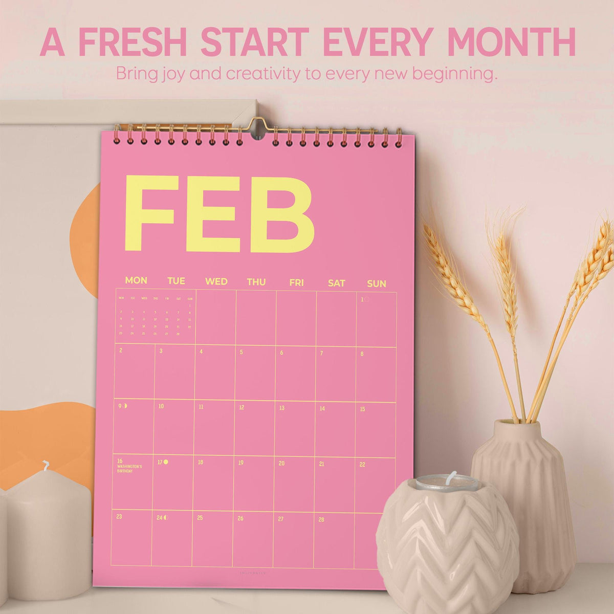 May include: A pink wall calendar for February with yellow text. The calendar is hanging on a wall and has a gold spiral binding. The text at the top says "A FRESH START EVERY MONTH" and "Bring joy and creativity to every new beginning."