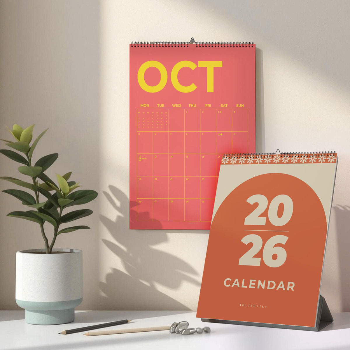 May include: A coral-colored wall calendar for October with yellow text and a matching desk calendar. The desk calendar has the numbers "2026" and the word "CALENDAR" in a rounded orange design. A potted plant and pencils are on the desk.