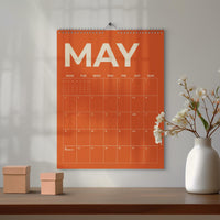 May include: Orange wall calendar for May with white text and grid. The calendar is hanging on a wall. A white vase with flowers and two small brown boxes are on a wooden surface.