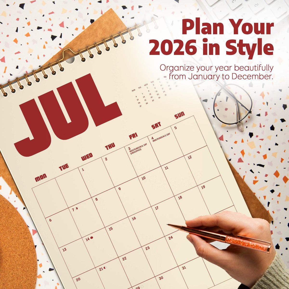 May include: A spiral-bound 2026 calendar open to July, with the word "JUL" in large red letters. The calendar is on a light beige background with a grid of dates. The text "Plan Your 2026 in Style" is at the top. A hand holds a pen.