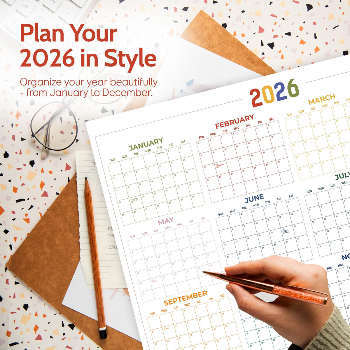 May include: A large 2026 wall calendar with the text "Plan Your 2026 in Style". The calendar is open to the months of January, February, March, May, June, July, and September. A hand holds a pen, pointing to a date. A pencil and glasses are also visible.
