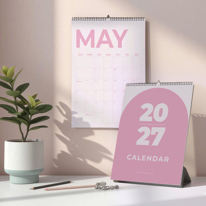 May include: A 2027 wall calendar with a pink arch design on the cover and a May monthly grid page hanging above it. The calendar features white text on a pink background, with a spiral binding at the top, displayed alongside a small potted plant.