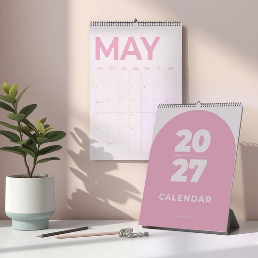 May include: A 2027 wall calendar with a pink arch design on the cover and a May monthly grid page hanging above it. The calendar features white text on a pink background, with a spiral binding at the top, displayed alongside a small potted plant.