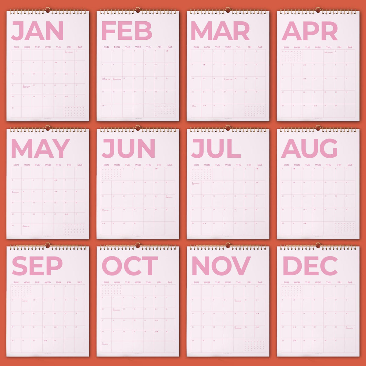 May include: A twelve-month wall calendar set featuring white pages with large, bold, light pink lettering for each month from January to December. Each page includes a grid layout for days of the week and dates, bound with a metal spiral at the top.