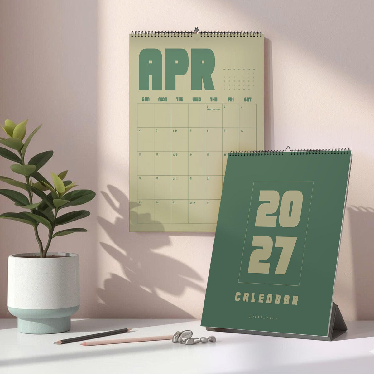 May include: A wall calendar showing the month of April in a light yellow hue and a dark green desk calendar displaying the year 2027. Both calendars feature wire-o binding and bold, block-style typography. A small potted plant sits to the left.