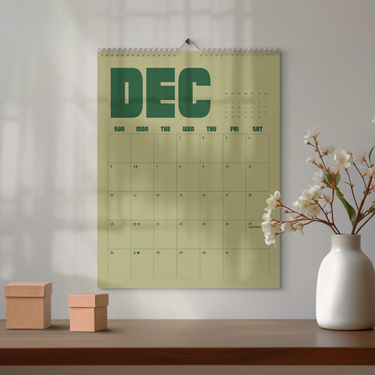 May include: A wall calendar for the month of December featuring a light olive green background with dark green bold text reading DEC. The grid displays days of the week and dates, with the 25th marked as Christmas and a small dot on the 27th.