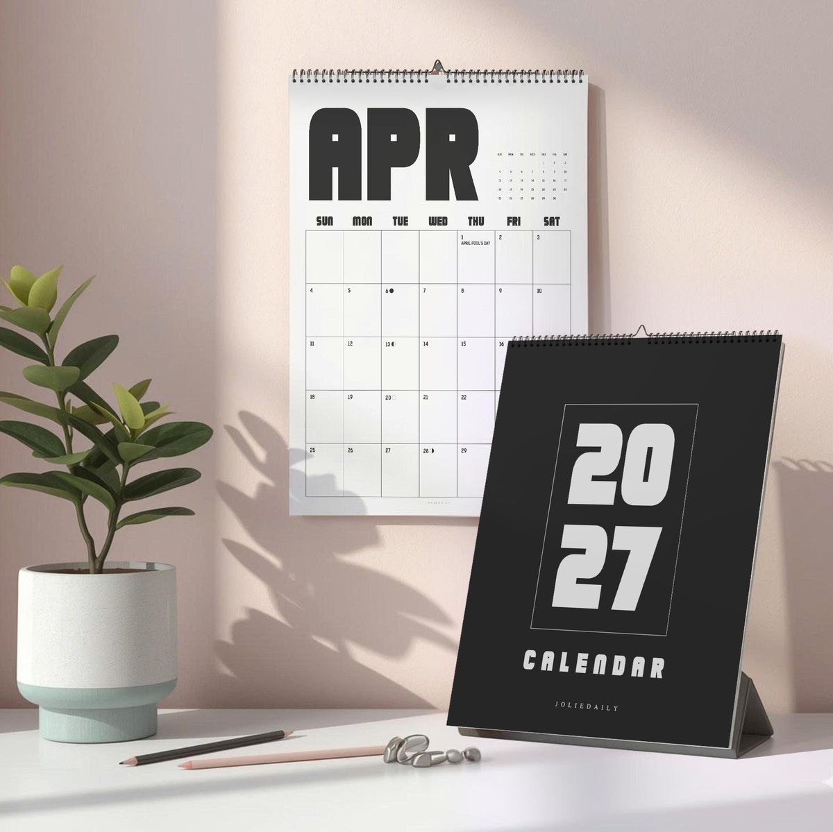 May include: A wall calendar showing the month of April with black text on a white background, and a standing desk calendar with a black cover featuring the white text 2027 CALENDAR. Both items are spiral-bound with a clean, modern design.