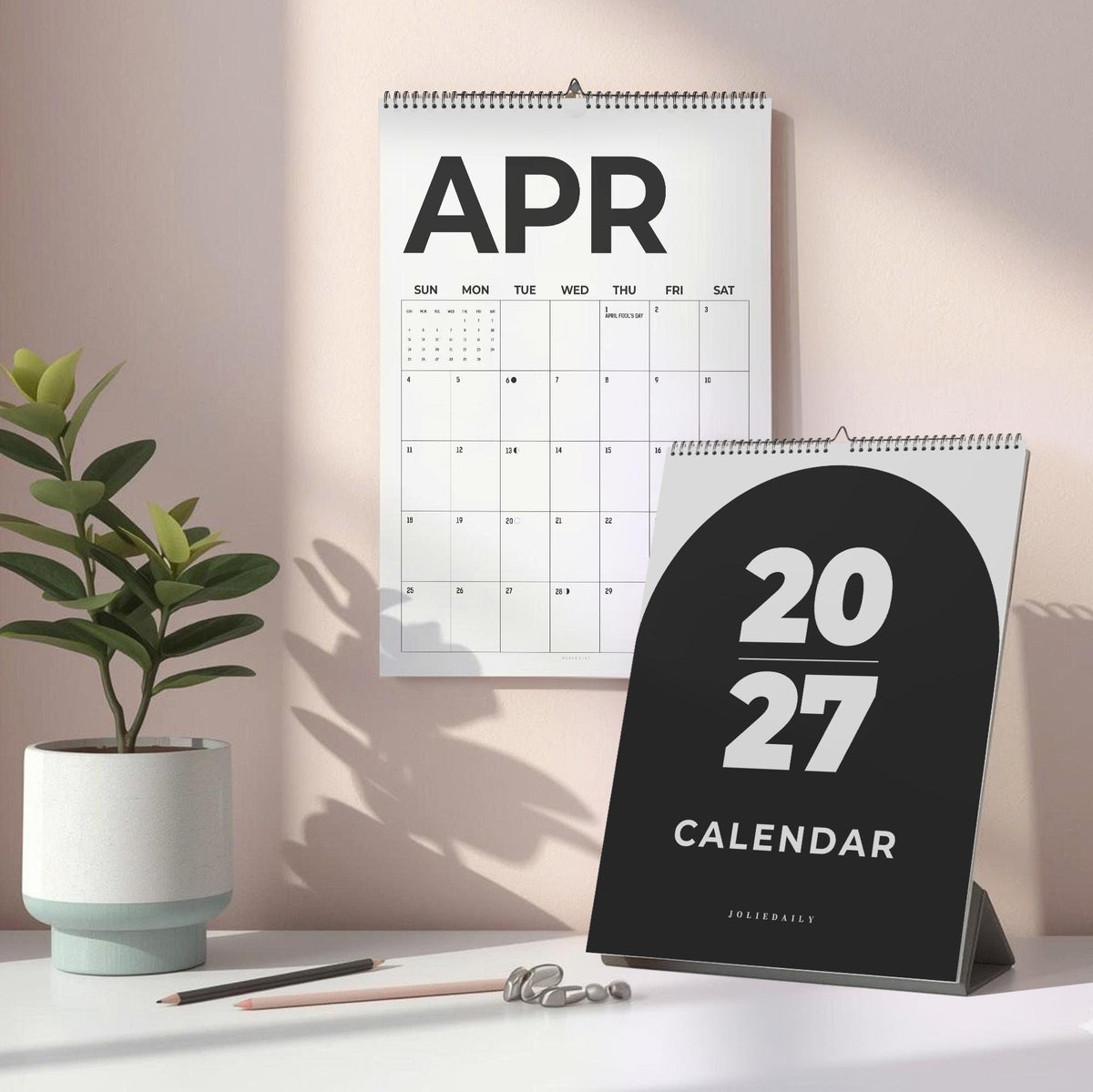 May include: A 2027 calendar with a black cover featuring white text and a wall-mounted calendar page showing the month of April. The calendar has a spiral binding at the top. The cover displays the year 2027 and the brand name JOLIEDAILY in white.