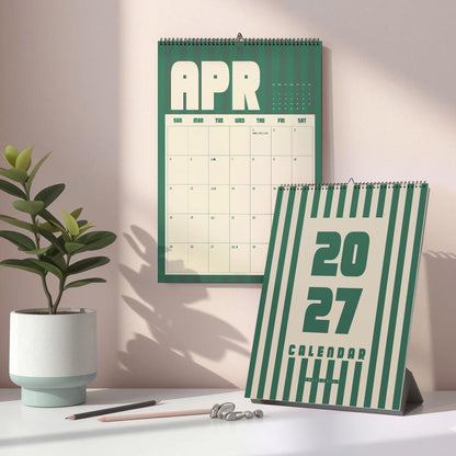 May include: A wall calendar displaying the month of April and a standing desk calendar featuring the year 2027. Both calendars have a dark green and cream striped design with bold typography. The desk calendar is positioned in front of the wall calendar.