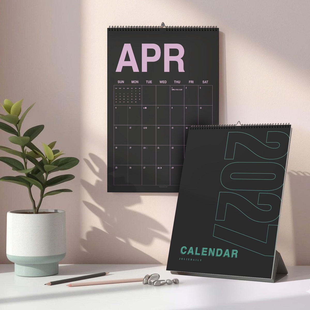 May include: A black wall calendar for April with large pink text and a black desk calendar for 2027 with teal text. The wall calendar features a grid layout for the month, while the desk calendar displays the year in a bold, vertical font.