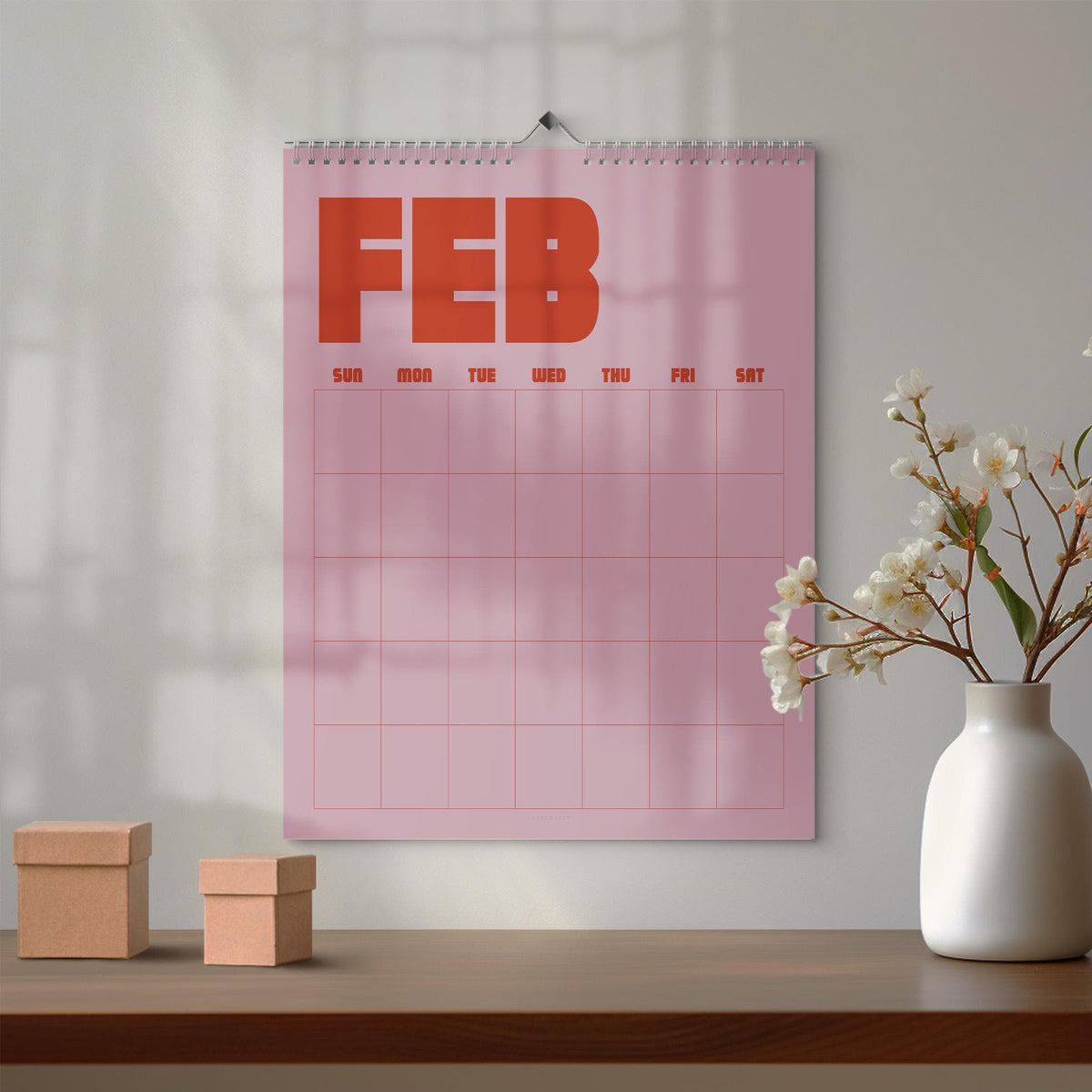 May include: A wall calendar with a pink background and the text FEB in large orange letters at the top. The calendar features a grid layout with days of the week labeled SUN, MON, TUE, WED, THU, FRI, and SAT in orange text above the empty grid squares.