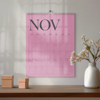 May include: A wall calendar with a pink background and the text NOV in large dark gray letters at the top. The calendar grid displays the days of the month with thin dark gray lines. It features a metal spiral binding at the top center.