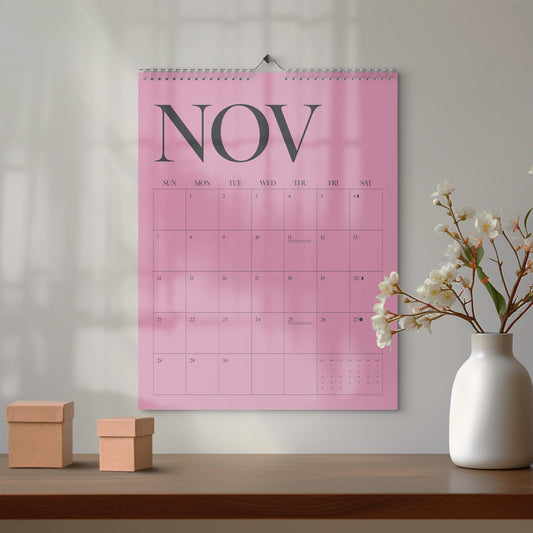 May include: A wall calendar with a pink background and the text NOV in large dark gray letters at the top. The calendar grid displays the days of the month with thin dark gray lines. It features a metal spiral binding at the top center.