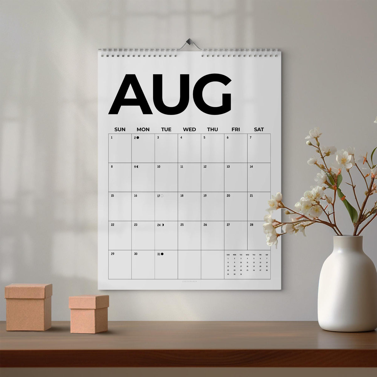 May include: A wall calendar featuring the month of August in large black sans-serif typography. The grid displays days of the week from Sunday to Saturday with numbered dates and small moon phase icons. It is bound with a silver metal spiral coil.