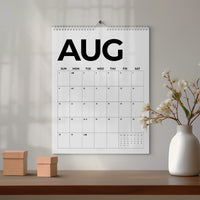 May include: A wall calendar featuring the month of August in large black sans-serif typography. The grid displays days of the week from Sunday to Saturday with numbered dates and small moon phase icons. It is bound with a silver metal spiral coil.