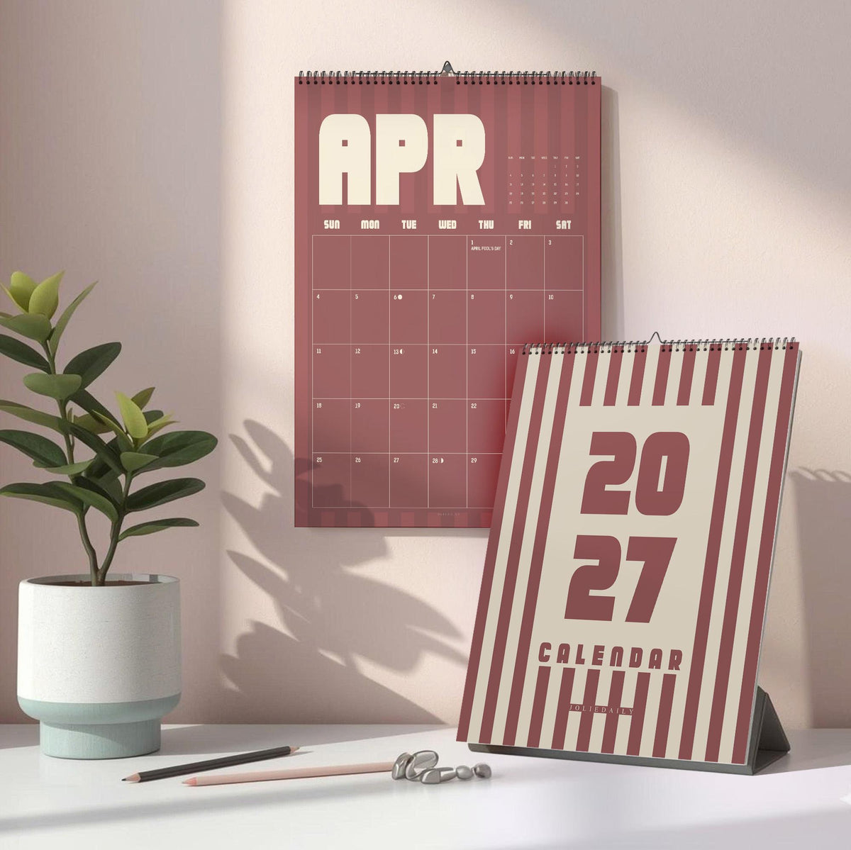 May include: A wall calendar showing the month of April with a dark red and cream striped design, and a matching desk calendar displaying the year 2027 in bold dark red lettering on a cream background with vertical dark red stripes.
