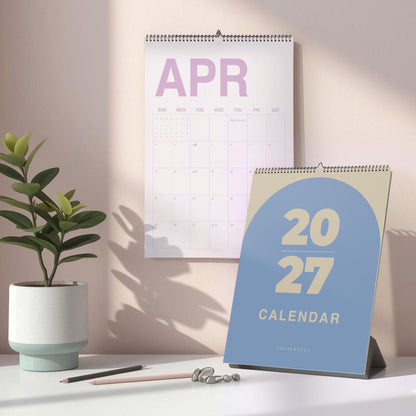 May include: A wall calendar showing the month of April with light purple text and a desk calendar featuring a blue arch design with the beige text 2027 CALENDAR. The wall calendar has a grid layout for days of the week and a metal spiral binding.