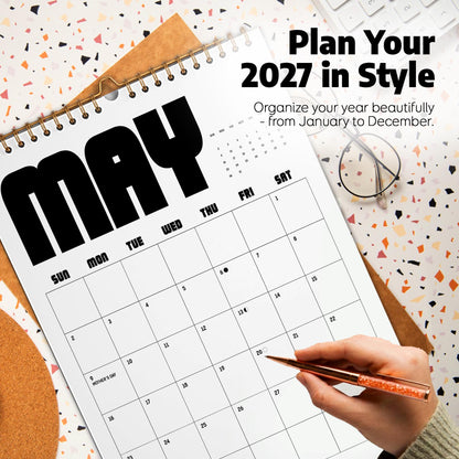 May include: A 2027 wall calendar with a white background and black text. The page shows the month of May with a grid of days, including the text Plan Your 2027 in Style and Organize your year beautifully - from January to December. A pen is present.