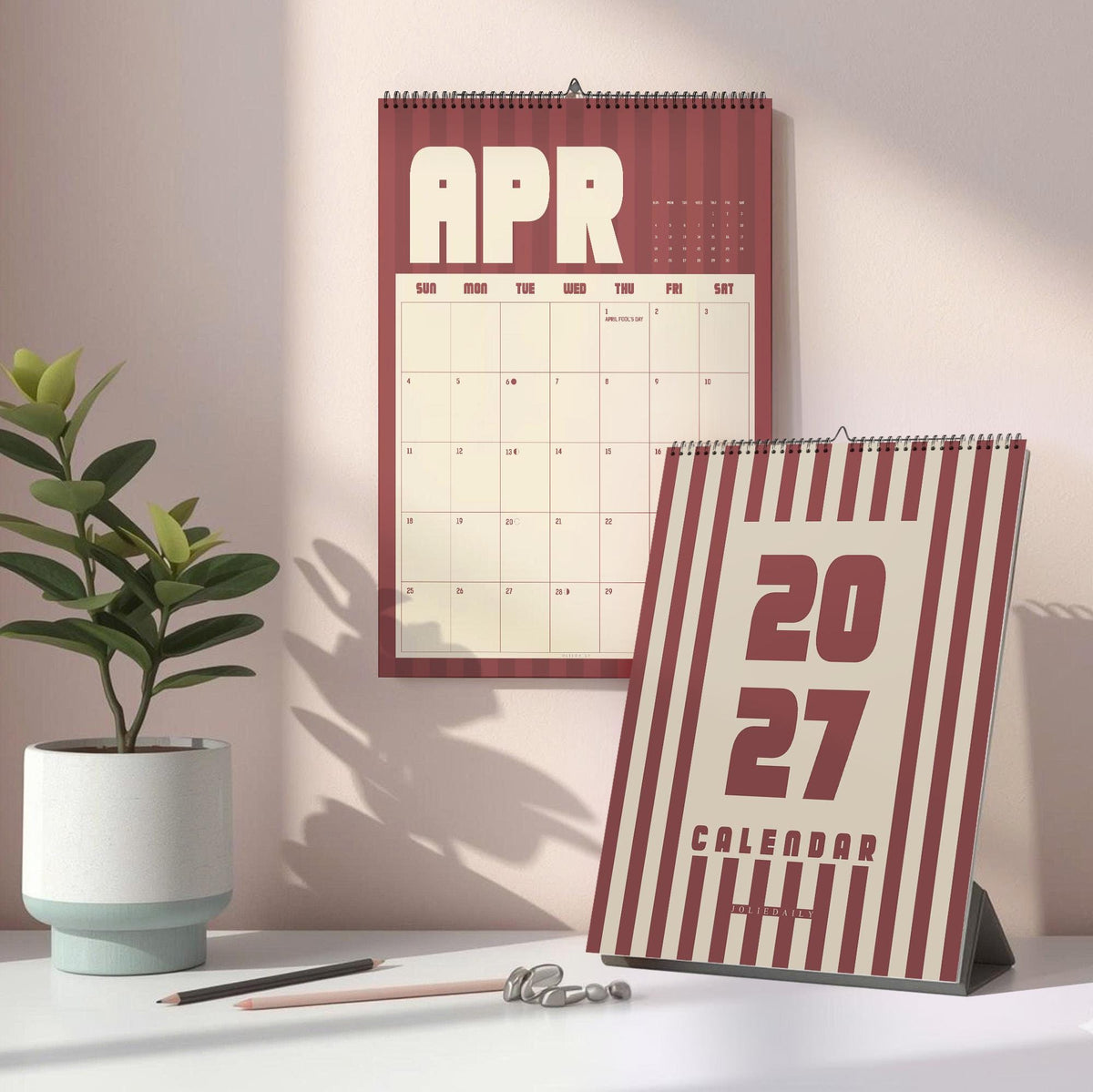 May include: A wall calendar showing the month of April and a standing desk calendar featuring the year 2027. Both calendars have a maroon and cream vertical striped pattern. The wall calendar displays a grid for dates, while the desk calendar shows 2027.