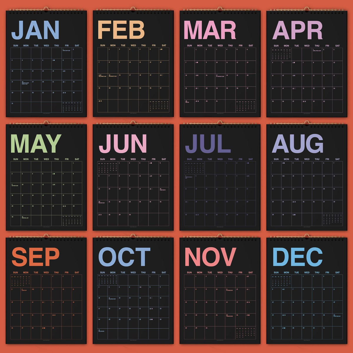 May include: A twelve-month wall calendar featuring black pages with white grid lines and colorful month names. Each month is displayed on a separate page with the abbreviation in large letters at the top, including JAN, FEB, MAR, APR, MAY, JUN, JUL, AUG, SEP, OCT, NOV, and DEC.