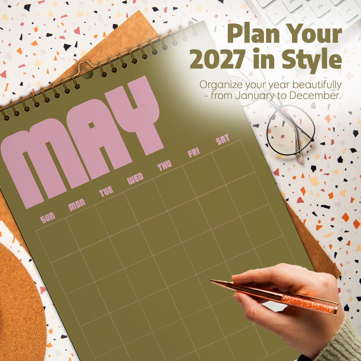 May include: A 2027 wall calendar with a spiral binding, featuring an olive green background and the month of May in large pink letters. The text reads Plan Your 2027 in Style and Organize your year beautifully - from January to December.