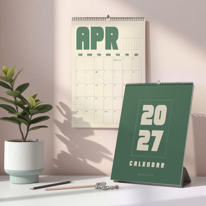 May include: A wall calendar for April 2027 with green text on a cream background, paired with a dark green desk calendar featuring the year 2027 in large cream font. Both items are spiral-bound and displayed with a potted plant and two pencils.