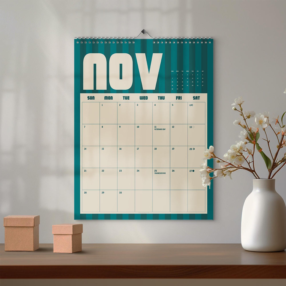 May include: A wall calendar for the month of November featuring a teal and dark teal vertical striped background. The calendar grid is cream-colored with black text, displaying the days of the week and dates, including Veterans Day and Thanksgiving.