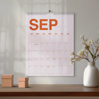 May include: A wall calendar for the month of September featuring large orange text reading SEP at the top. The grid displays days of the week from Sunday to Saturday with numbered dates in a light orange color, suspended by a silver metal wire coil.