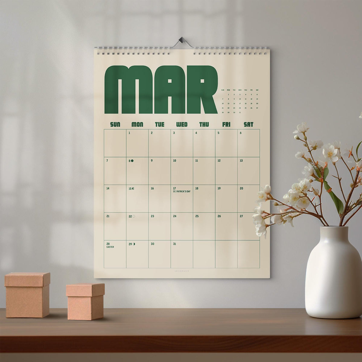 May include: A wall calendar for the month of March featuring large dark green block lettering at the top. The calendar grid is printed in dark green on a cream background, displaying days of the week and dates, including St. Patrick's Day.