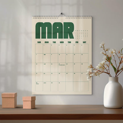 May include: A wall calendar for the month of March featuring large dark green block lettering at the top. The calendar grid is printed in dark green on a cream background, displaying days of the week and dates, including St. Patrick's Day.