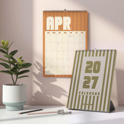 May include: A wall calendar showing the month of April with a vertical orange striped background and a desk calendar featuring the year 2027 in large bold font with olive green vertical stripes on a white background.