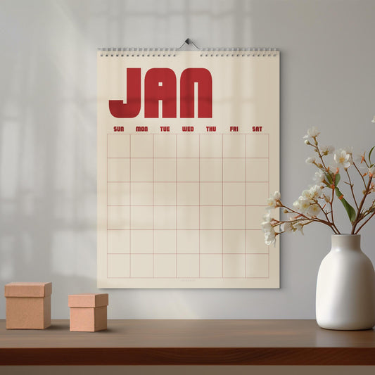 May include: A wall calendar with a cream-colored background and a bold red header reading JAN. The calendar features a grid layout for the days of the week, labeled SUN, MON, TUE, WED, THU, FRI, and SAT in red text, suspended by a silver wire coil.