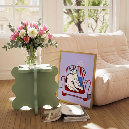 May include: A framed cat art print featuring a black and white cat lounging in a red armchair. A bouquet of pink tulips and white dahlias sits in a clear vase on a light green side table. A stack of books rests on the floor.