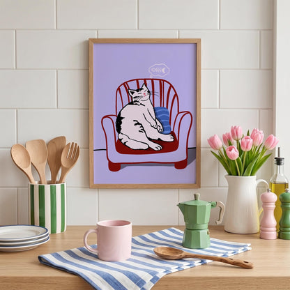 May include: A framed art print featuring a relaxed cat sitting in a red armchair with a blue pillow. The cat is white with black markings, and a thought bubble with a fishbone is above its head. The background is a light purple.