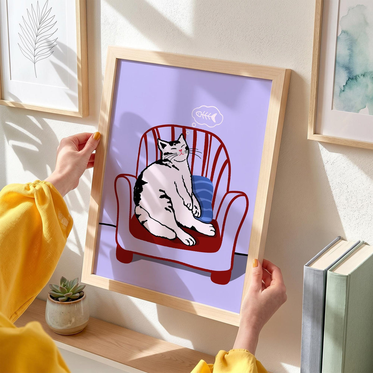 May include: A framed art print featuring a cartoon cat relaxing in a red armchair with a blue pillow. The cat is white with black markings. A thought bubble above the cat shows a fishbone. The background is a light purple.