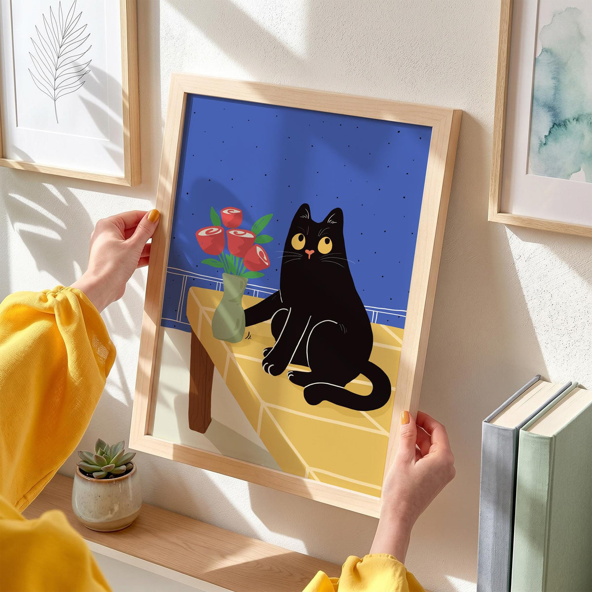 May include: A framed art print featuring a black cat sitting on a yellow table with a vase of red roses. The background is blue. The print is in a light wood frame.